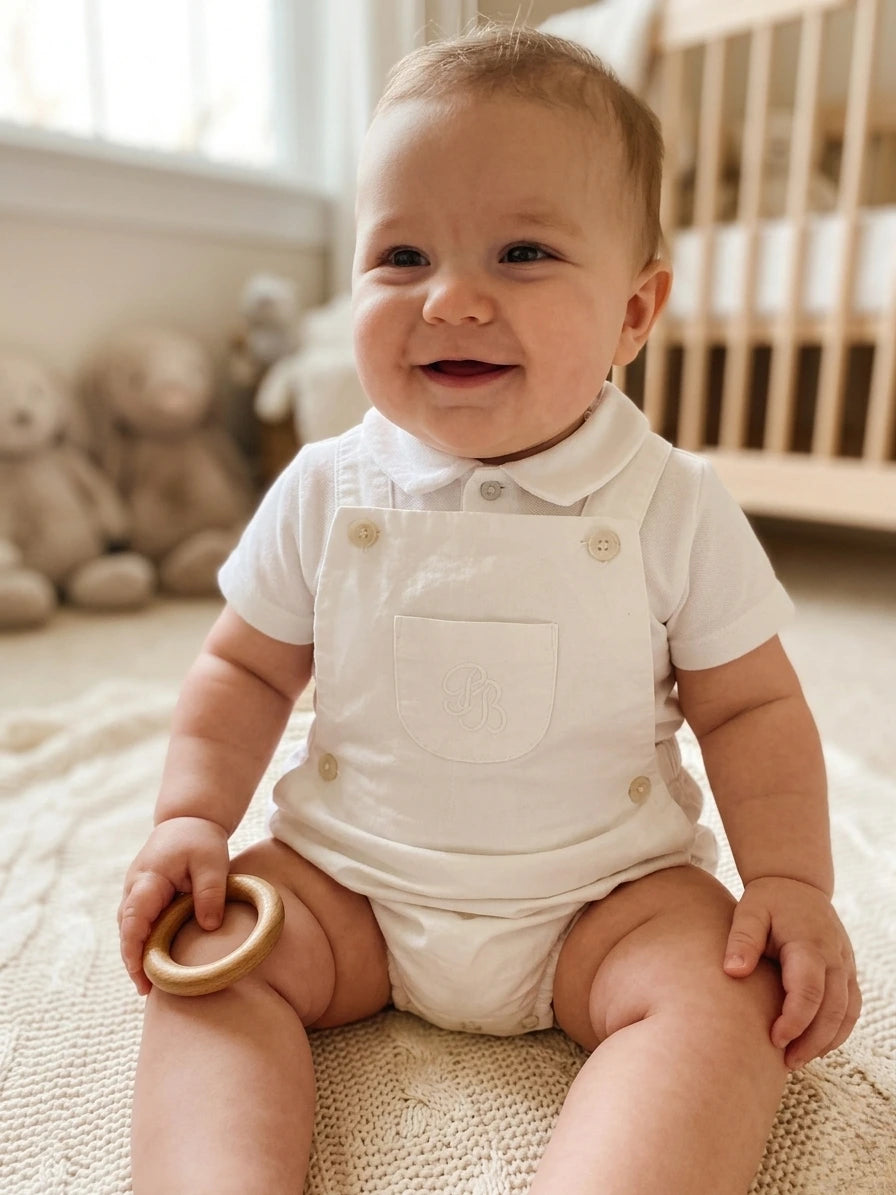 Baby in a white outfit sitting on a rug with teddy bears in the background