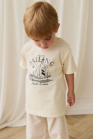 Child wearing a cream t-shirt with a sailboat graphic and text, standing against a white curtain.