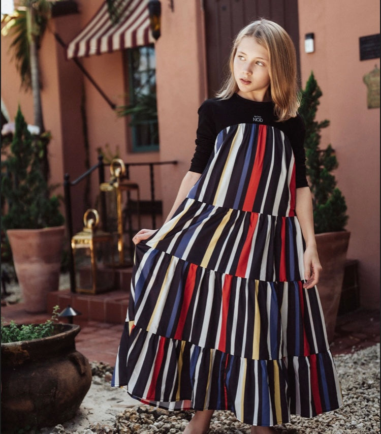 Young blonde girl in a vertically striped tiered maxi dress with a black long-sleeved top, outdoors.
