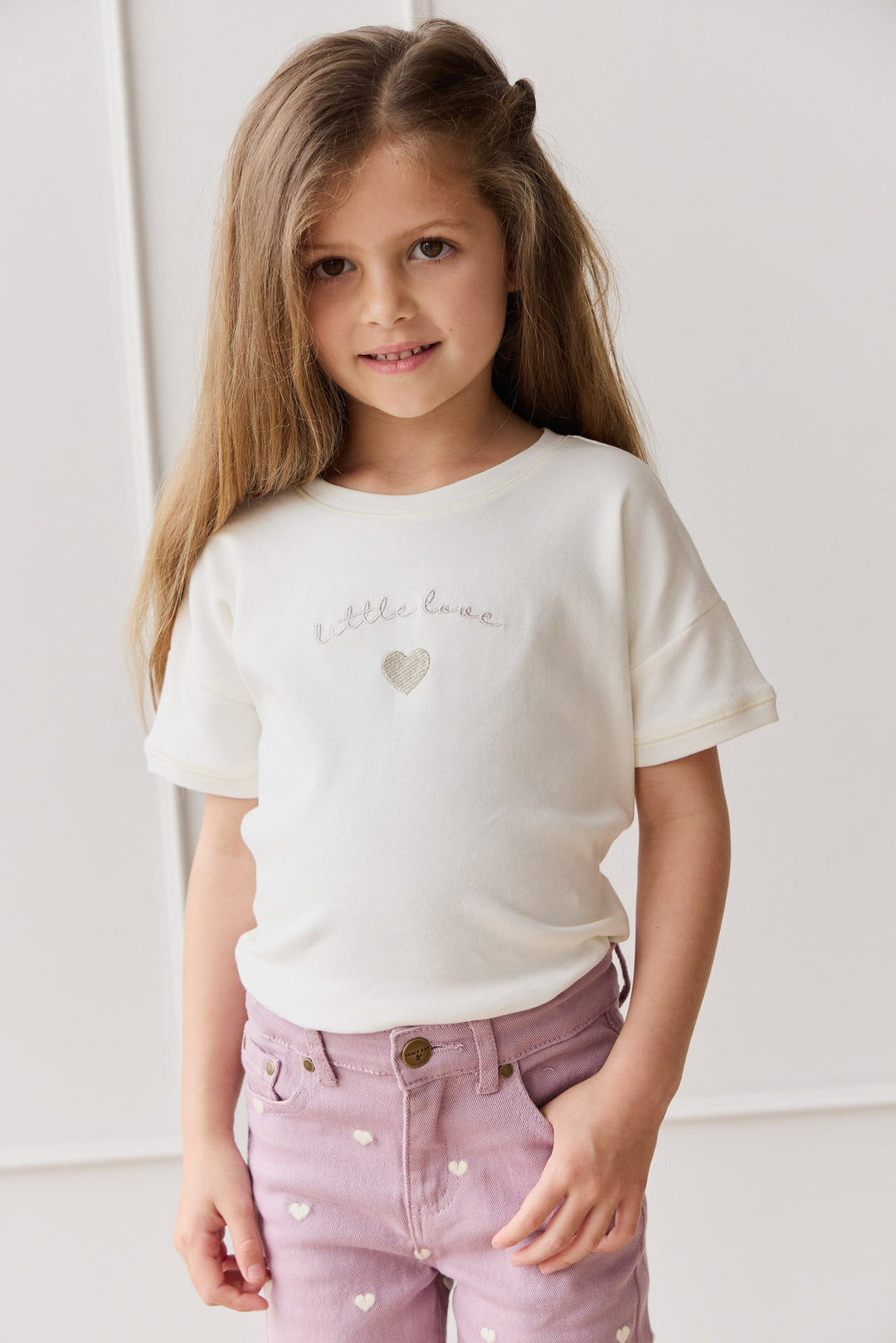 She is wearing a short-sleeved, off-white t-shirt. On the front of the t-shirt, slightly off-center, is the handwritten cursive text "little love" in a delicate, light gray or silver color.