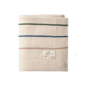 A folded cream-colored knitted throw blanket with thin horizontal stripes in green, blue, and pink, featuring a woven label that reads 'OMANI HOME BEAUTY'.