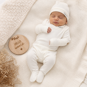 Newborn baby in a white outfit and hat lying on a soft surface with a 'Hello World' sign.