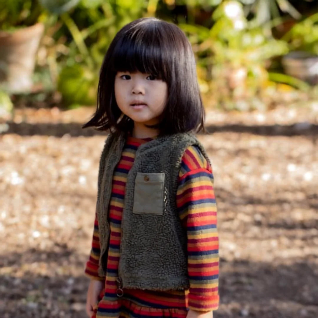 Child wearing a striped shirt and vest standing outdoors with a blurred natural background