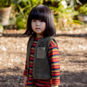 Child wearing a striped shirt and vest standing outdoors with a blurred natural background