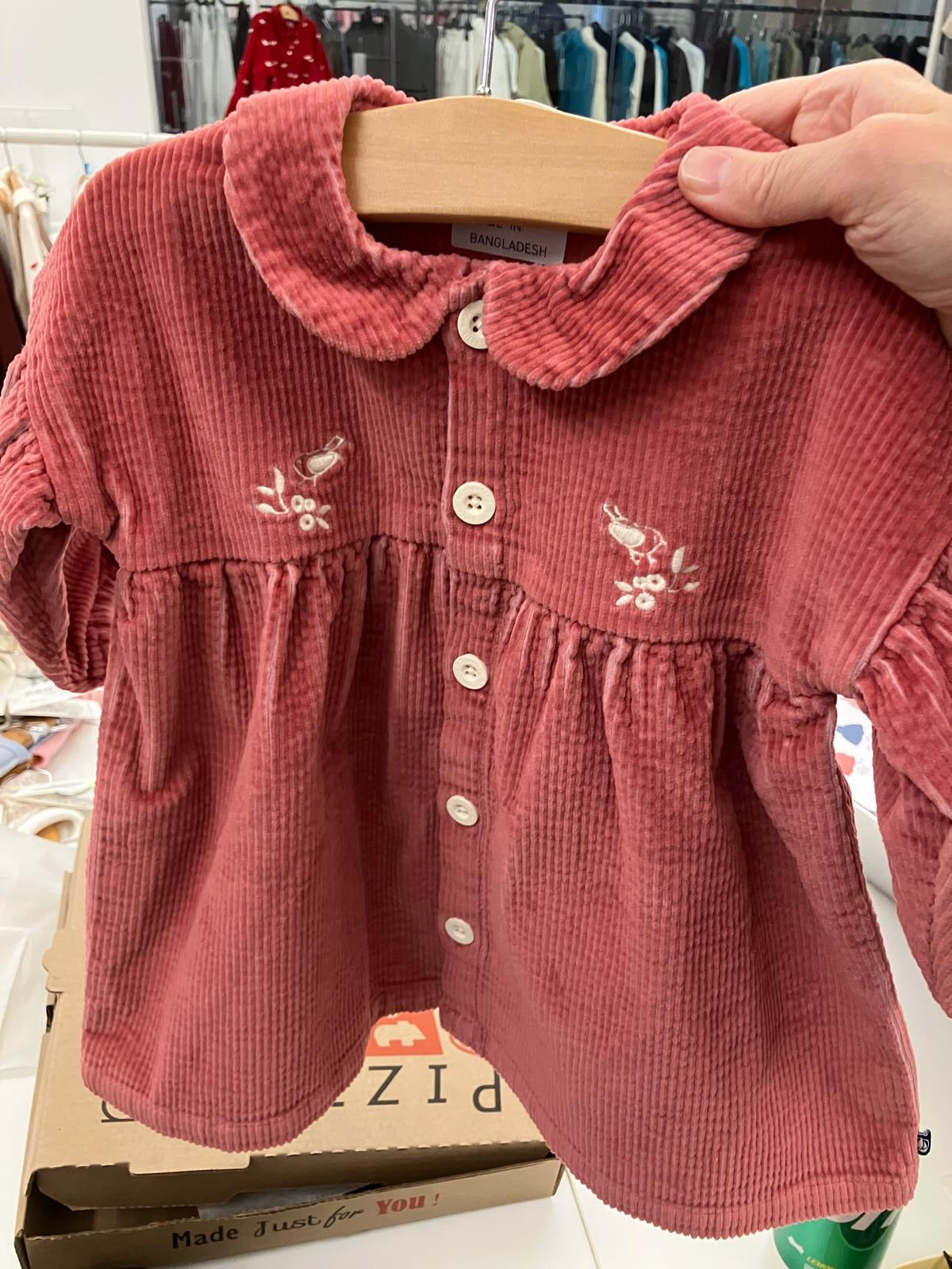 Red corduroy dress with floral embroidery on a hanger, with a blurred store background.