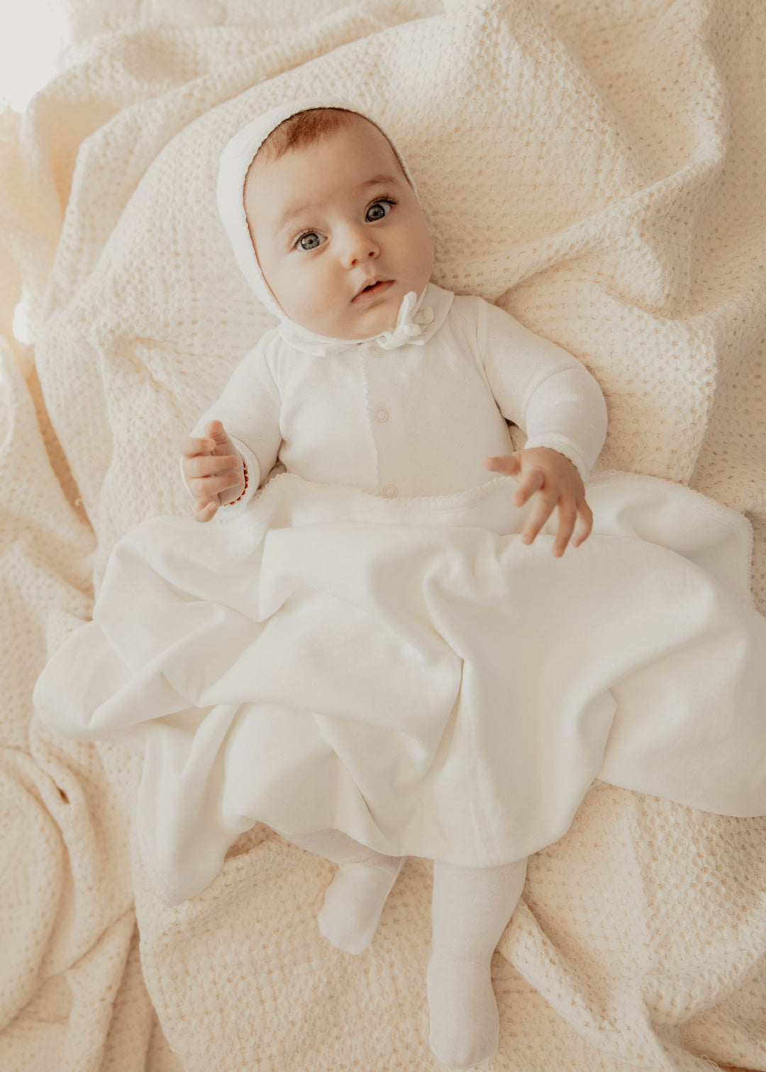 Baby in a white outfit lying on a soft beige blanket