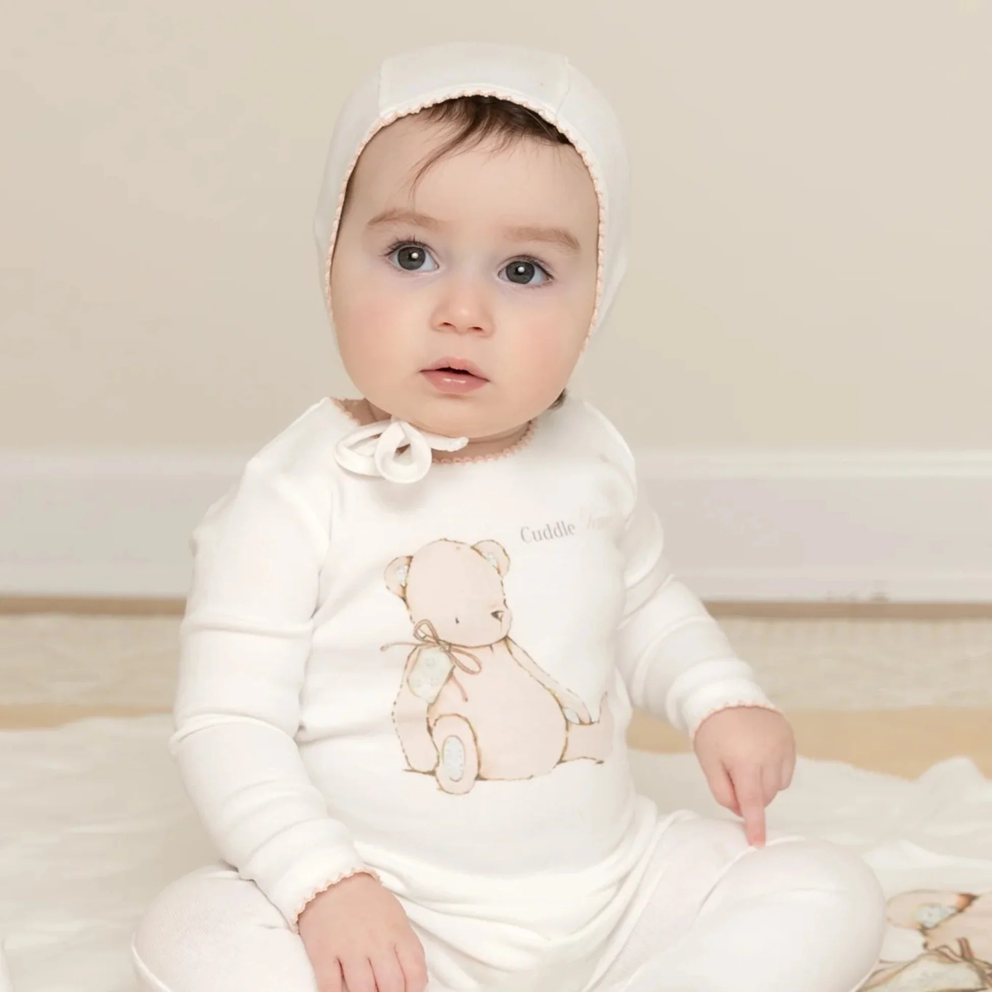 Baby wearing a white outfit with a teddy bear design on a light-colored floor.