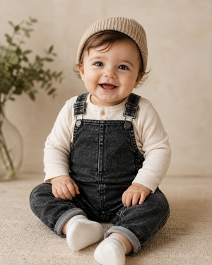 A smiling baby sitting on a textured beige floor, wearing a cream ribbed long-sleeve top, dark gray denim overall, a beige knit beanie, and white socks.