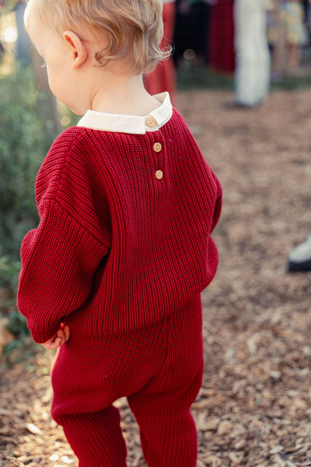 Child wearing a red knitted outfit with a white shirt underneath, standing outdoors.
