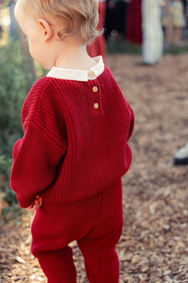 Child wearing a red knitted outfit with a white shirt underneath, standing outdoors.