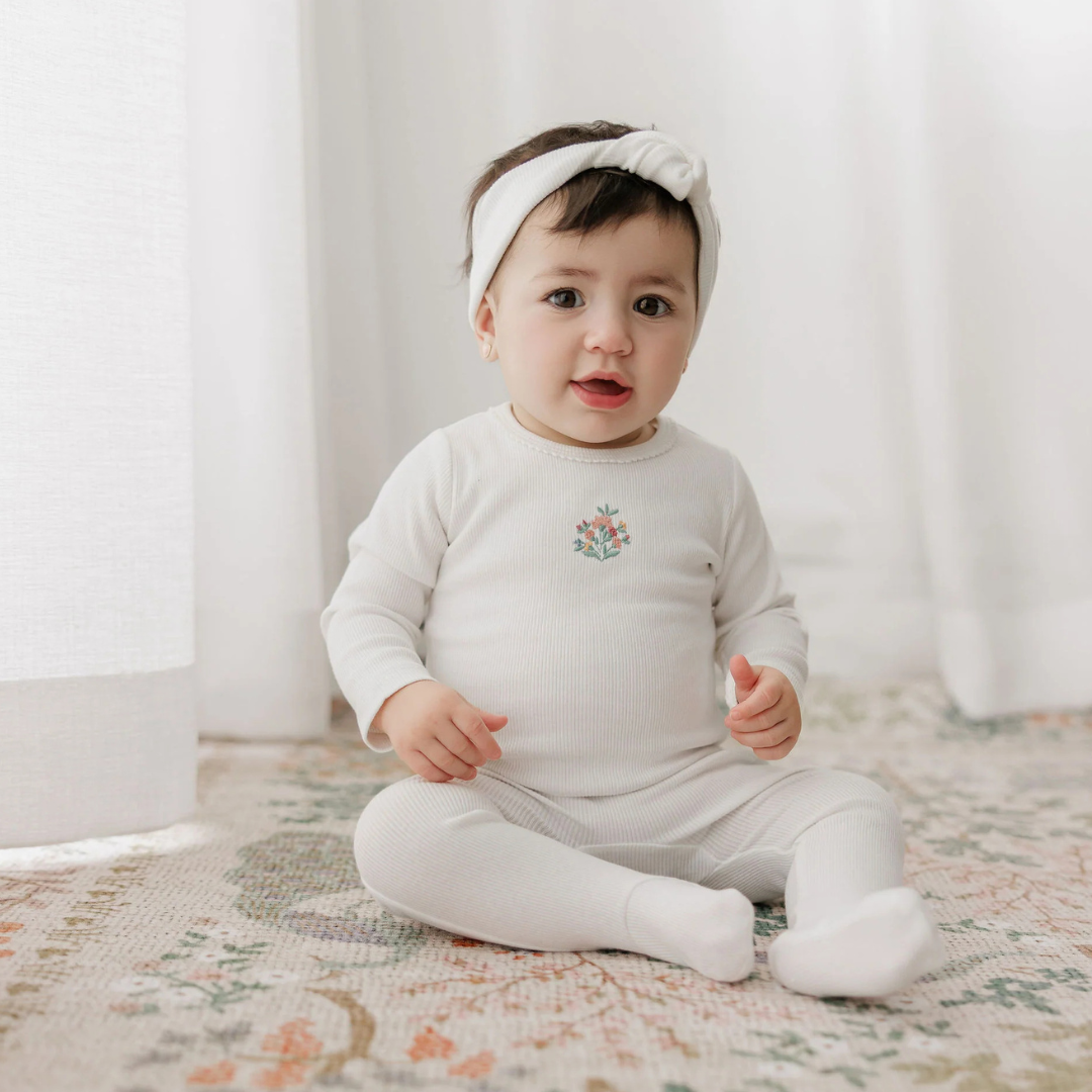 Baby wearing a white outfit with a floral design, sitting on a patterned rug.
