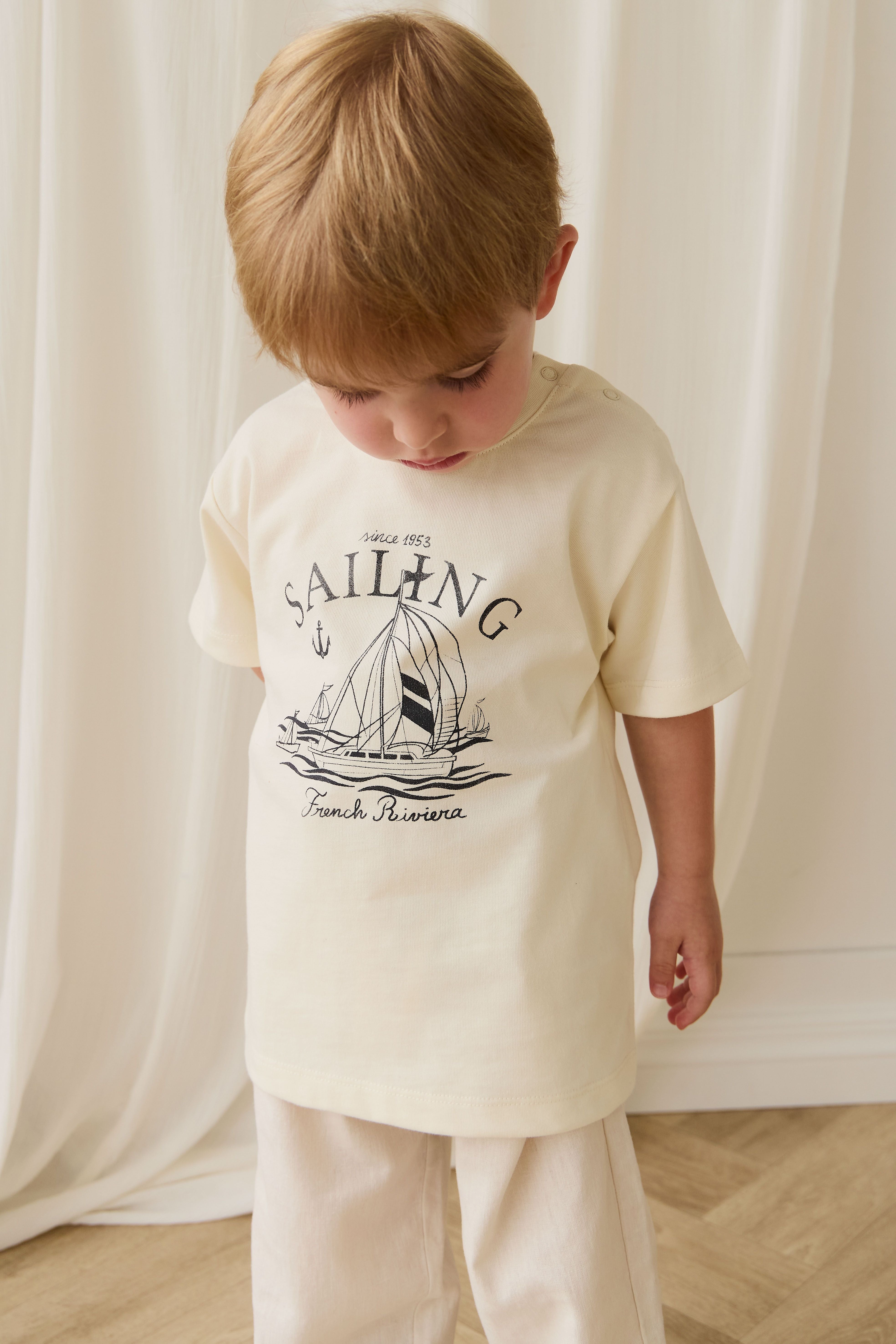 Child wearing a cream t-shirt with a sailboat graphic and text, standing against a white curtain.