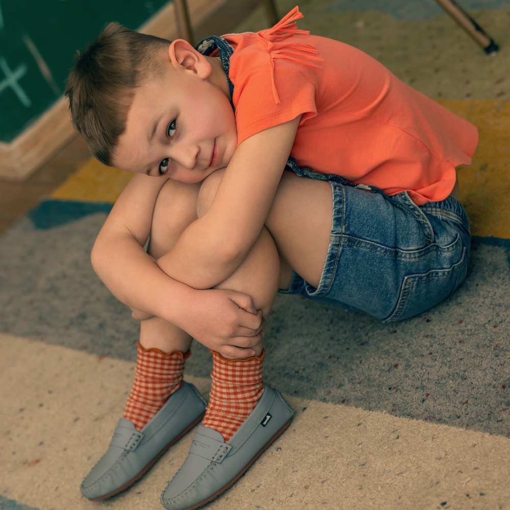 Child sitting on a colorful floor wearing an orange shirt, denim shorts, and checkered socks.