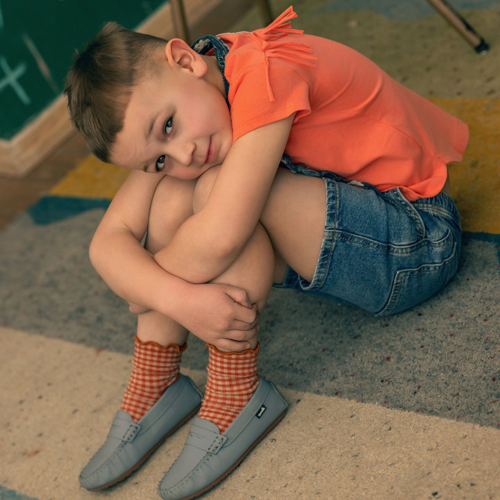 Child sitting on a colorful floor wearing an orange shirt, denim shorts, and checkered socks.