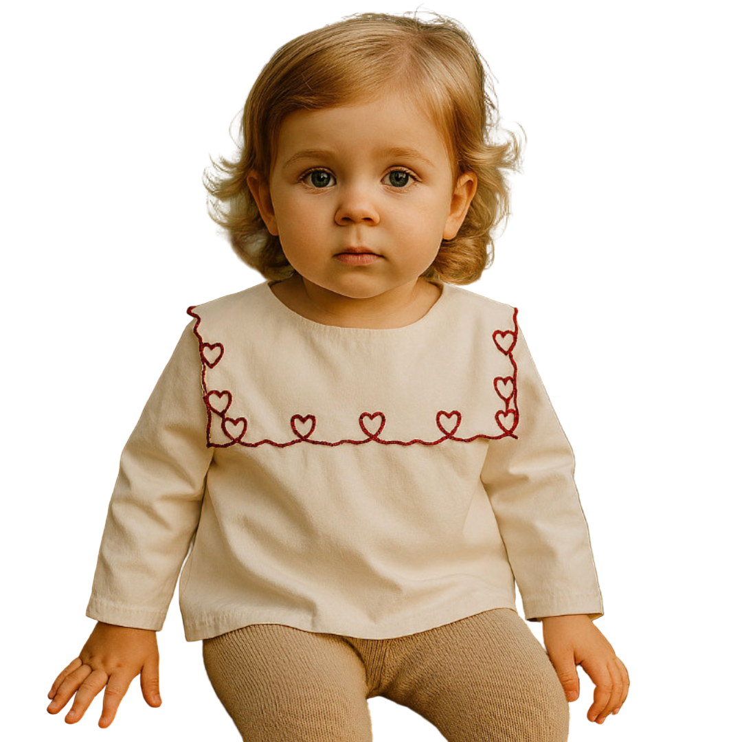 Child wearing a cream-colored top with red heart patterns, sitting outdoors.
