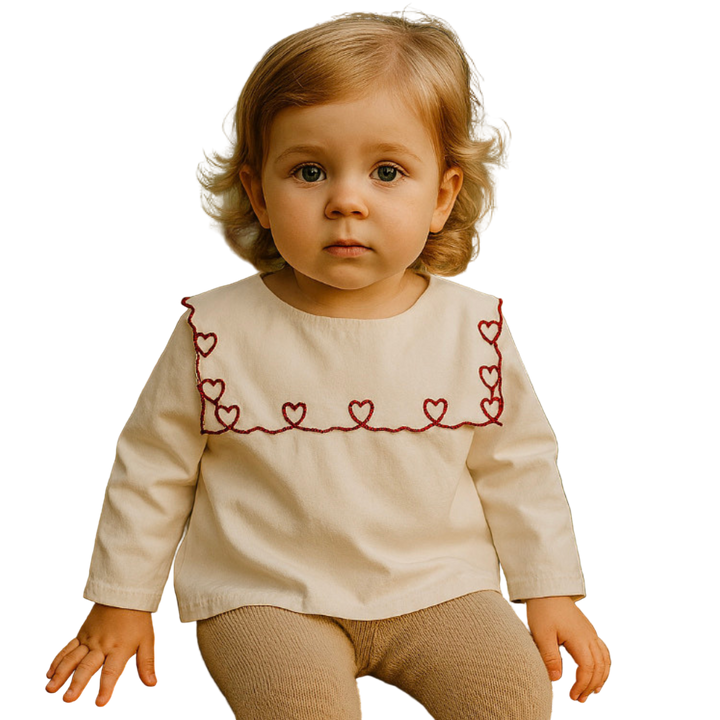 Child wearing a cream-colored top with red heart patterns, sitting outdoors.