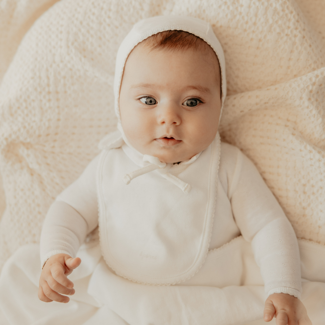 Baby wearing a white outfit and bonnet sitting on a soft beige surface