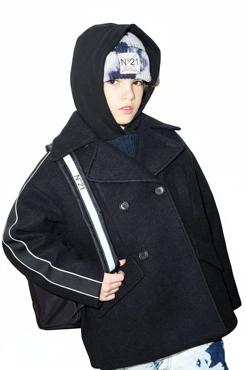 Person wearing a black coat with a hood, holding a white bag, against a light-colored wall.