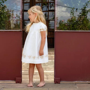 Young girl in a white dress standing in front of a red wall with plants.