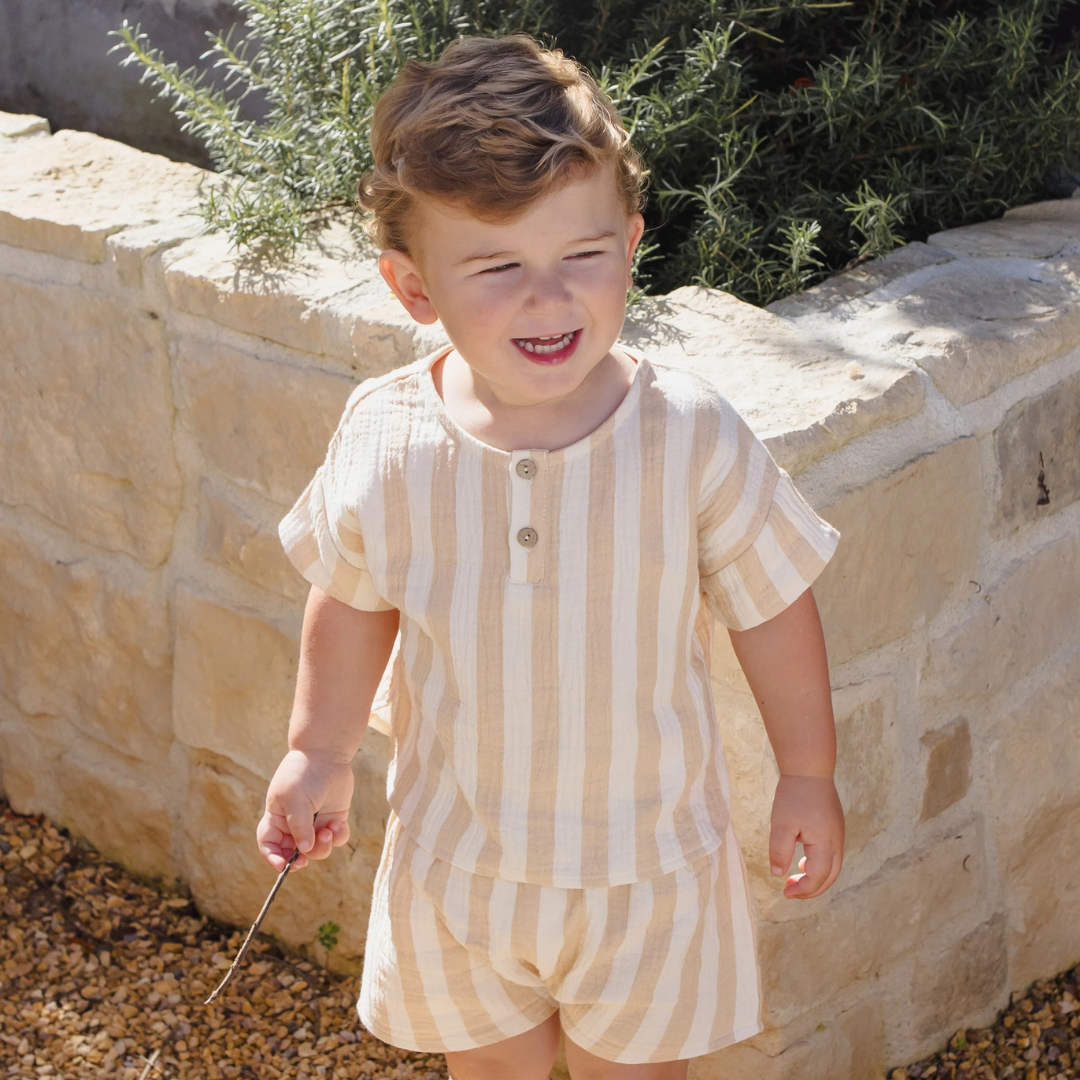 Child wearing a striped outfit standing in front of a stone wall with greenery.