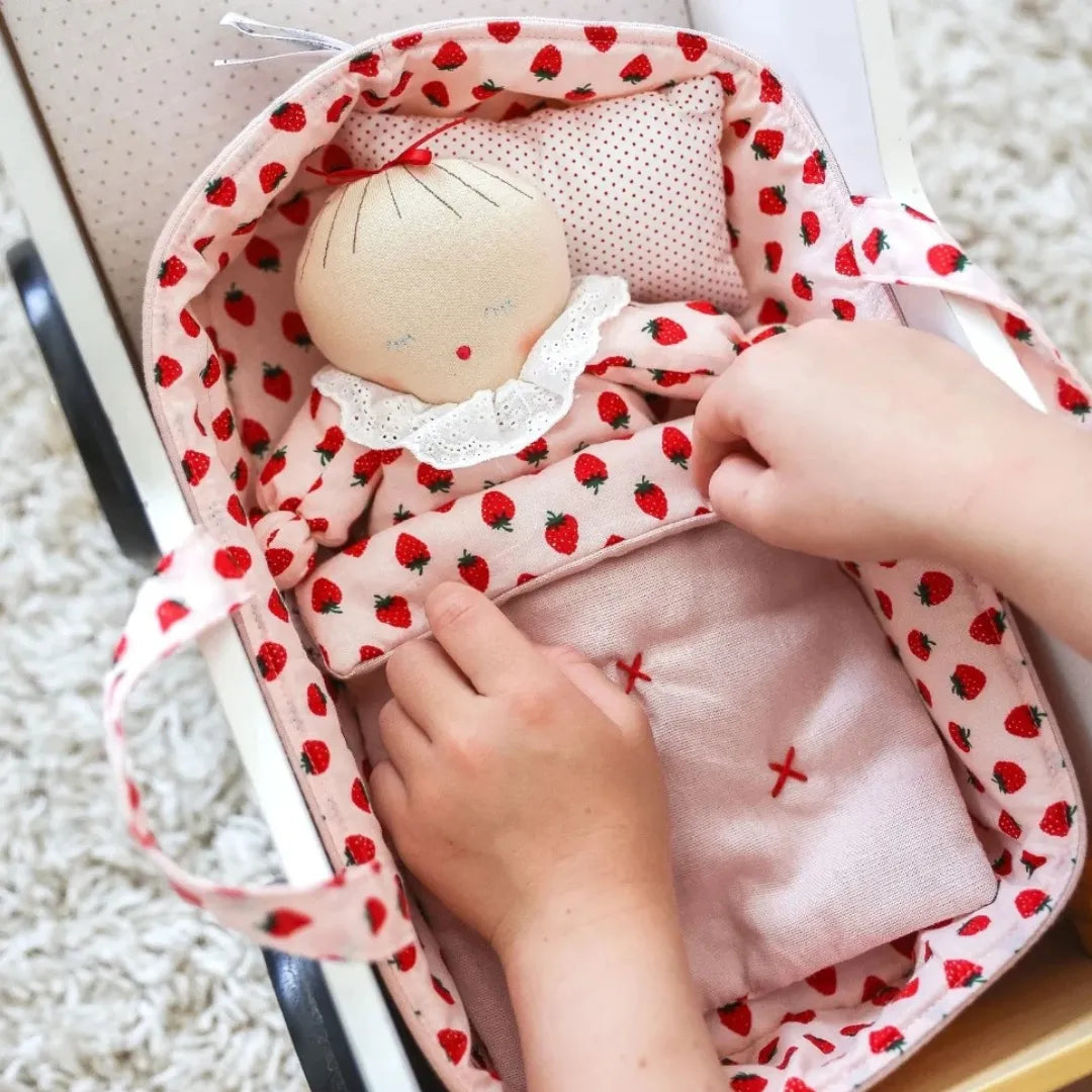 A baby doll playset inside a fabric bassinet, featuring a cream-colored doll with a strawberry-patterned bonnet and a matching pink and red strawberry-print bassinet with a baby blanket and pillow, shown with hands interacting with it.