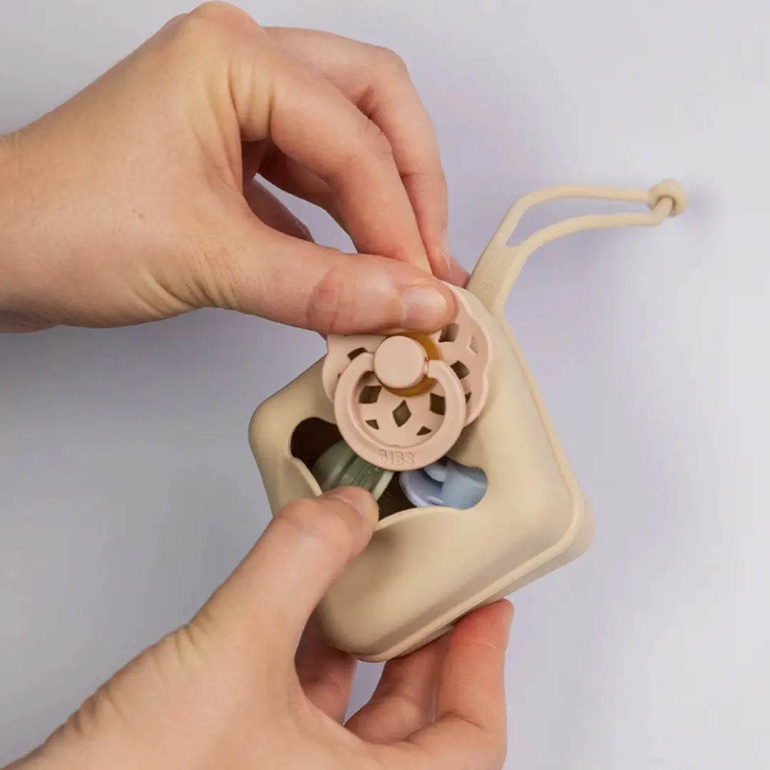 Hand holding a beige pacifier case with a pink pacifier inside against a light gray background