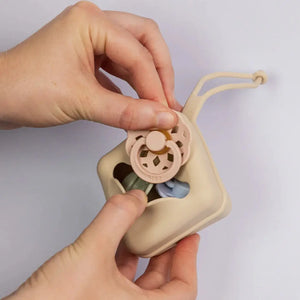 Hand holding a beige pacifier case with a pink pacifier inside against a light gray background