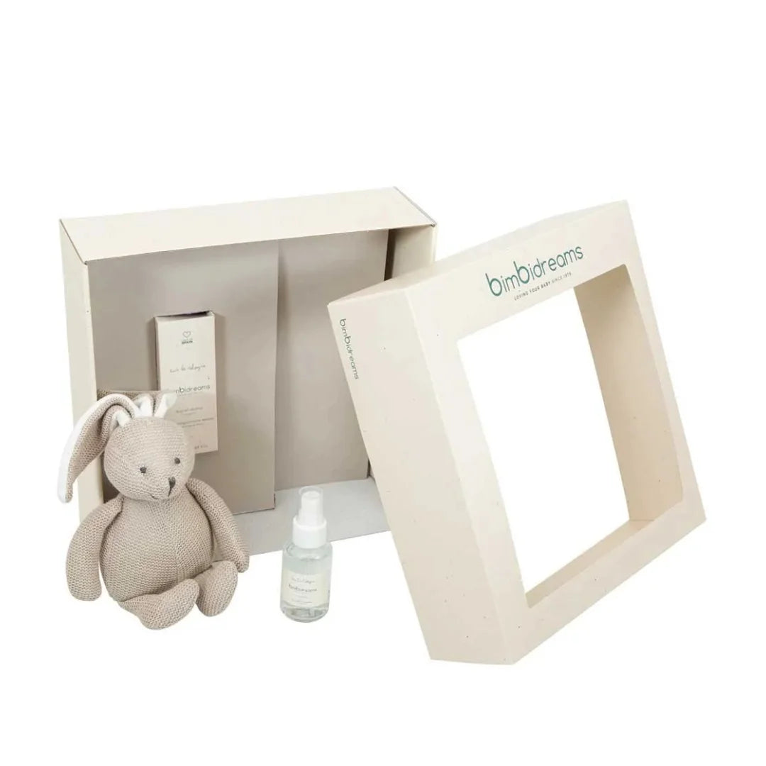 A baby gift set featuring a beige plush bunny, a small clear bottle with a white dropper cap, and a cream-colored gift box with a windowed lid. The brand name 'bimbr dreams' is printed on the lid.