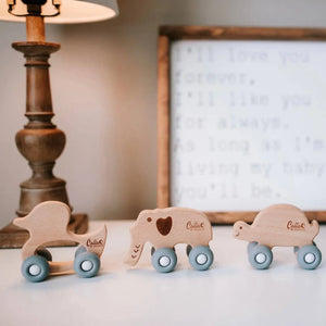 Three wooden animal toys on a surface with a lamp and framed picture in the background.