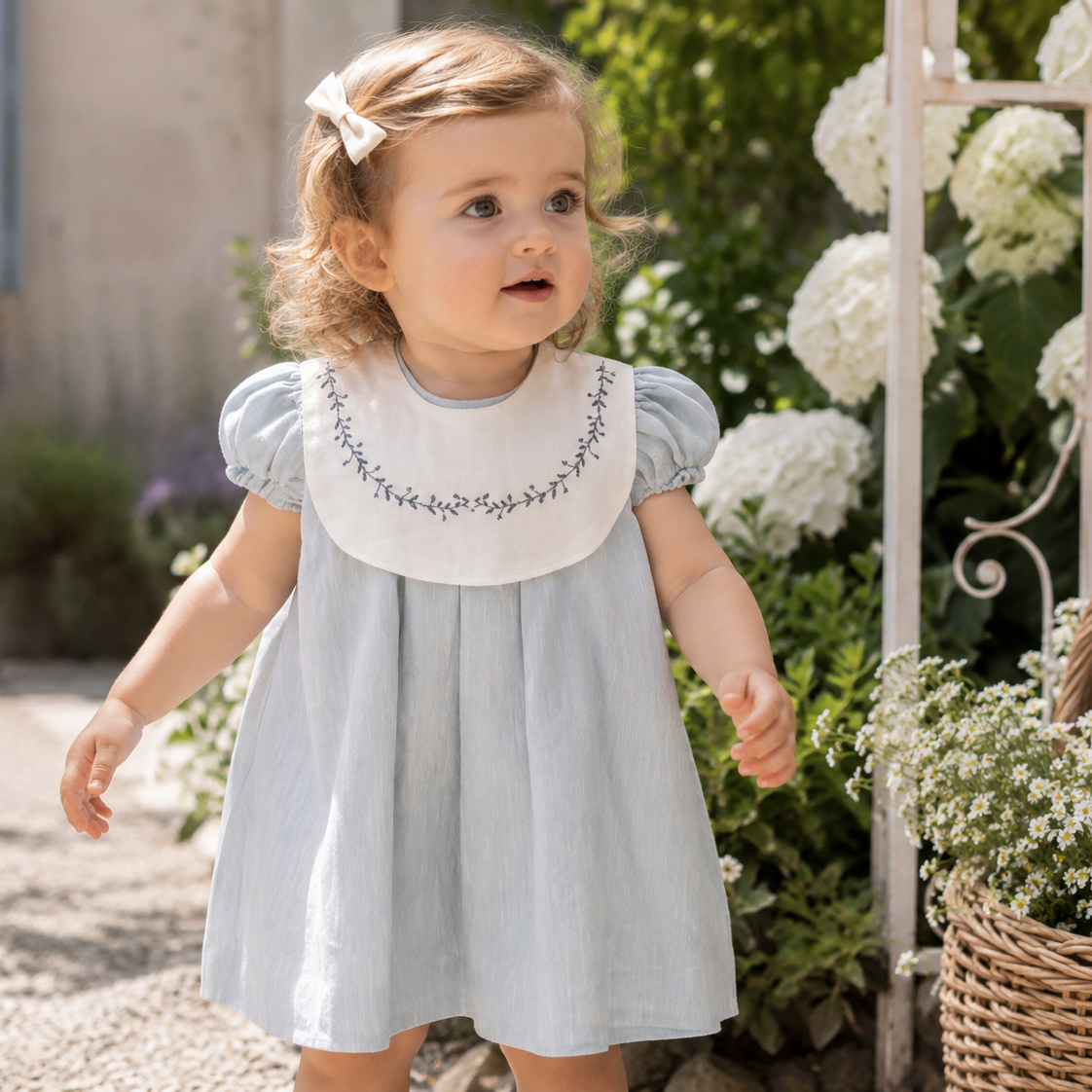 Young girl in a light blue dress with floral embroidery standing outdoors among flowers.