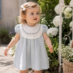Young girl in a light blue dress with floral embroidery standing outdoors among flowers.