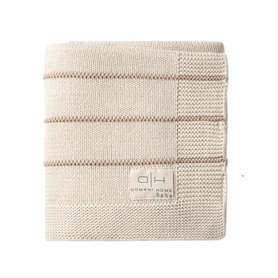 A folded cream-colored knit blanket with horizontal stripes and a square patch on the right side, featuring a small woven label that reads 'DOMANI HOME Bebe'.