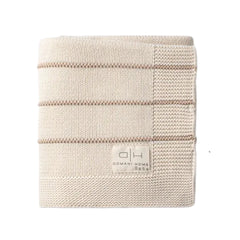 A folded cream-colored knit blanket with horizontal stripes and a square patch on the right side, featuring a small woven label that reads 'DOMANI HOME Bebe'.
