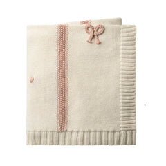 A folded cream-colored knit blanket featuring a vertical dusty pink textured stripe, a matching pink knotted bow detail, and a ribbed border, shown against a plain white background.