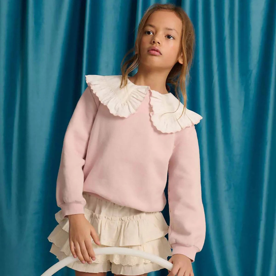A young girl wearing the pink Gingersnaps sweater with a ruffled collar in a park.