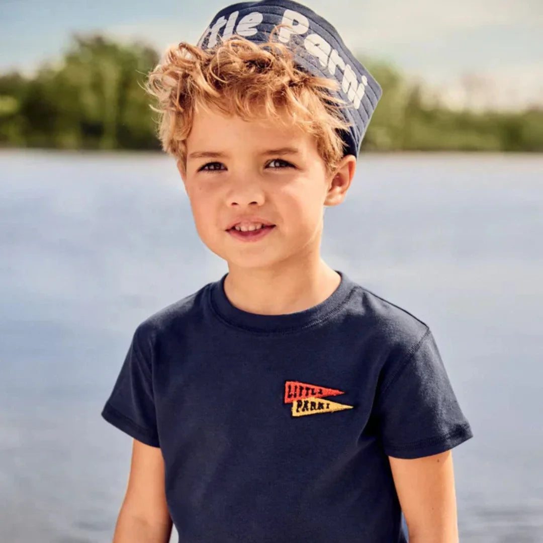 Young boy wearing a navy t-shirt with a logo and cap, standing by a body of water.