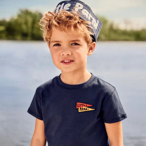Young boy wearing a navy t-shirt with a logo and cap, standing by a body of water.