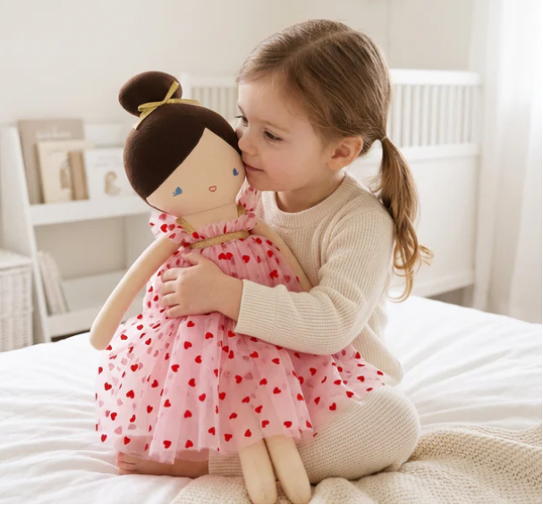 Child hugging a doll with a pink dress on a bed in a bright room.