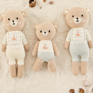 Three knitted teddy bear dolls wearing cream and beige outfits, arranged on a textured beige fabric background with small seashell props.